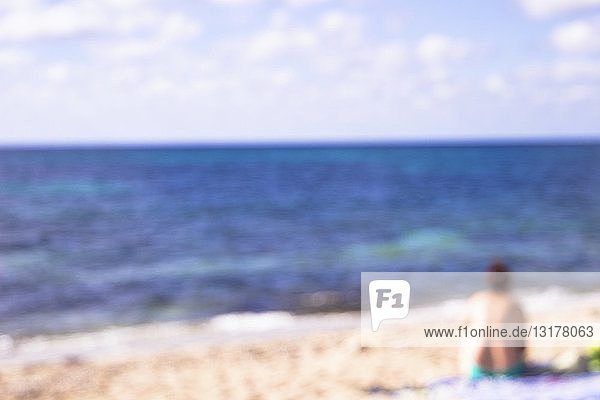 Unscharfe Rückenansicht einer Frau  die am Strand sitzt und auf die Aussicht schaut