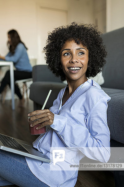 Happy woman with soft drink sitting on floor using laptop