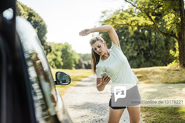 Sportliche junge Frau  die sich beim Autofahren auf einem Parkplatz streckt und ihr Handy benutzt