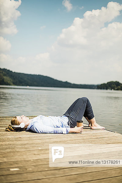 Woman lying on jetty at a lake with headphones and takeaway coffee
