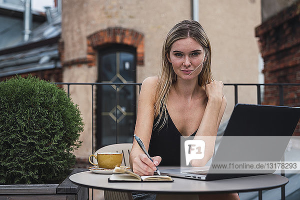 Porträt einer jungen Frau  die mit Laptop auf dem Balkon sitzt und Notizen macht