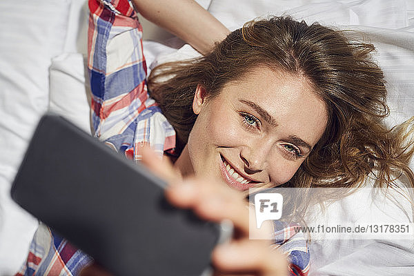 Porträt einer lachenden Frau  die auf dem Bett liegt und sich mit einem Smartphone Selbsthilfe nimmt