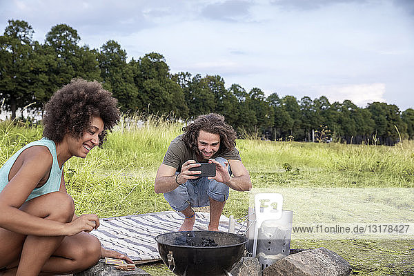 Lächelnder junger Mann mit Freundin beim Fotografieren eines Barbecue-Grills in der Natur