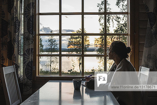 Finnland  Lappland  junge Frau sitzt am Fenster und schaut auf einen See