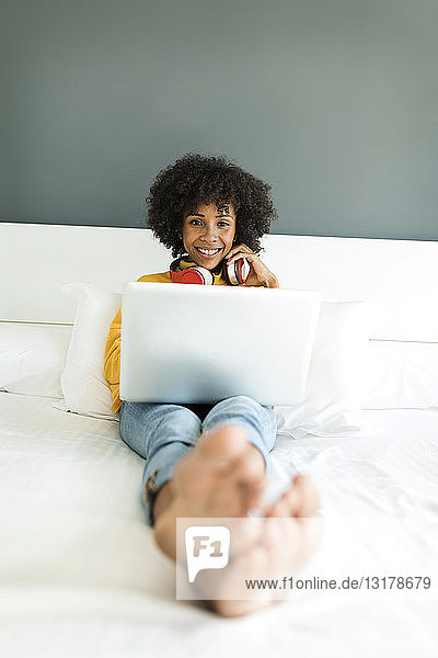 Happy woman lying on bed with headphones and laptop