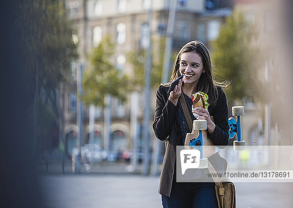 Smiling young woman with longboard and snack in the city using cell phone