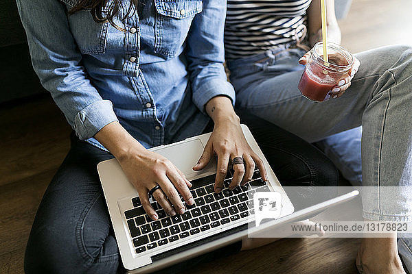 Close-up of two young women sitting on floor with soft drink and laptop