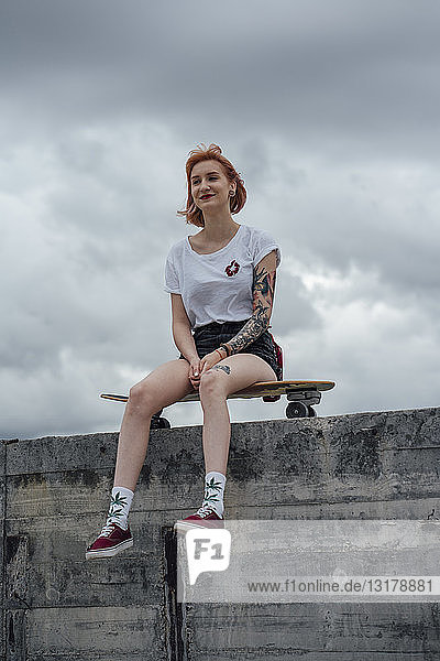 Lächelnde junge Frau sitzt auf einem Skateboard auf einer Betonwand