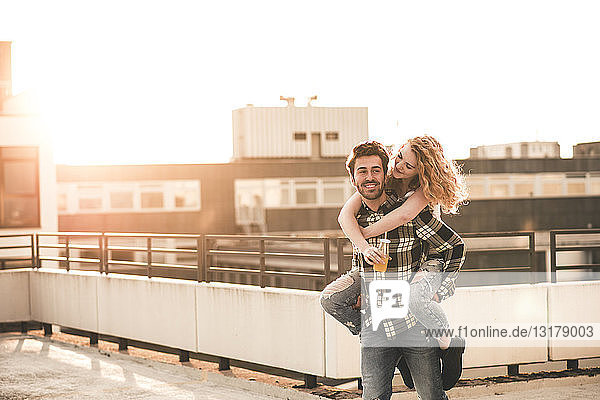 Porträt eines jungen Mannes  der seine Freundin bei Sonnenuntergang huckepack auf der Dachterrasse mitnimmt