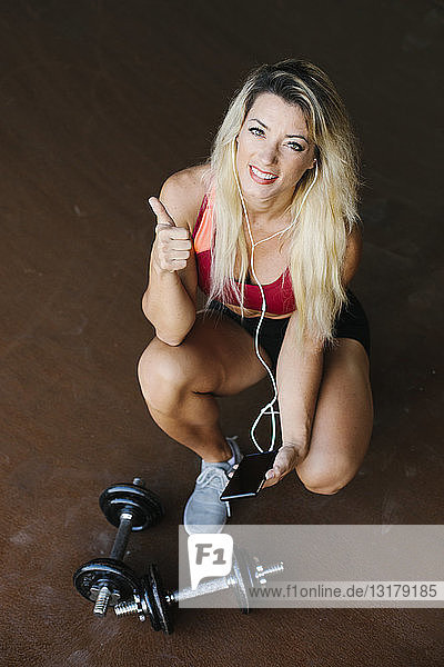 Porträt einer lächelnden athletischen Frau mit Kurzhanteln  Mobiltelefon und Kopfhörern