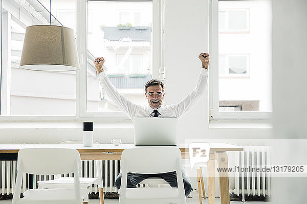Laughing businessman with laptop on table cheering in office