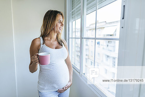 Schwangere Frau schaut aus dem Fenster und hält einen Becher