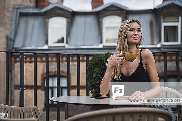Porträt einer blonden jungen Frau  die auf der Dachterrasse sitzt und eine Tasse Kaffee trinkt