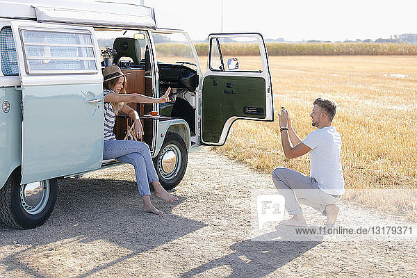 Young man taking cell phone picture of girlfriend inside camper van in rural landscape