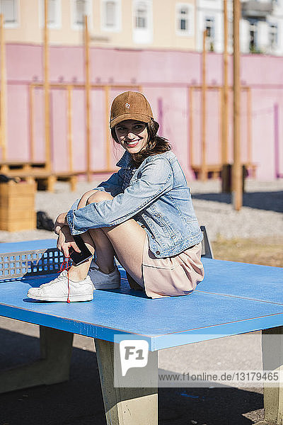Lächelnde junge Frau mit Handy auf Tischtennisplatte sitzend