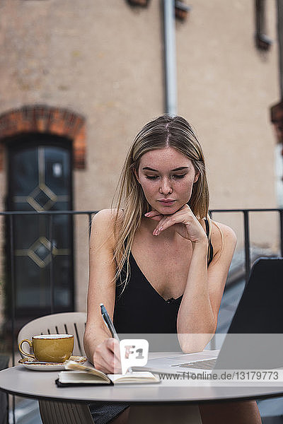 Portrait of young woman sitting on balcony with laptop taking notes