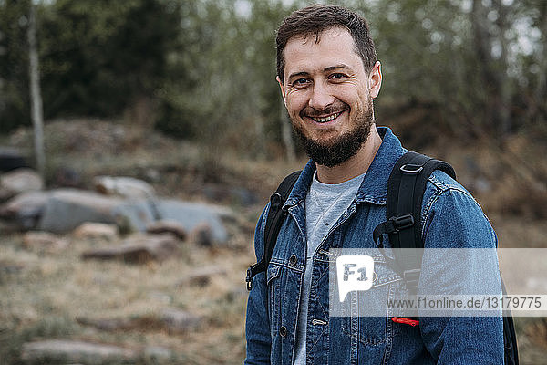 Porträt eines lächelnden Mannes mit Rucksack und Jeansjacke in Natur