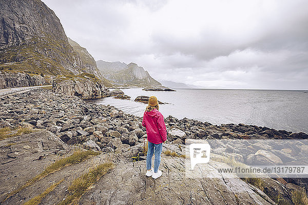 Norway  Lofoten  back view of young woman wearing rainjacket standing on a rock looking at distance