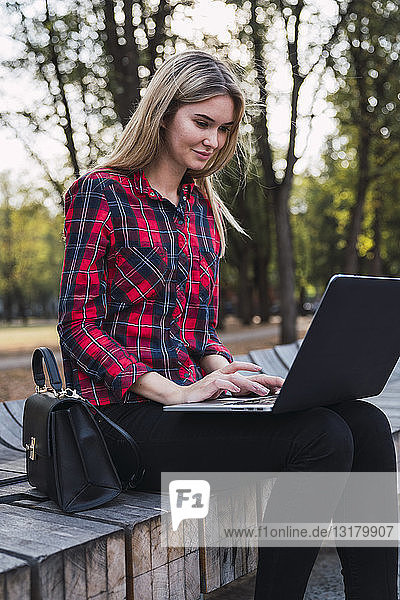 Modische junge Frau  die im Freien auf einer Bank sitzt und am Laptop arbeitet
