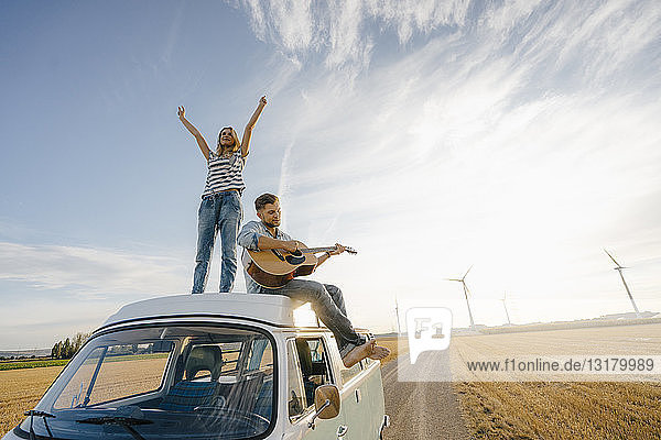 Glückliches Paar mit Gitarre auf dem Dach eines Wohnmobils in ländlicher Landschaft