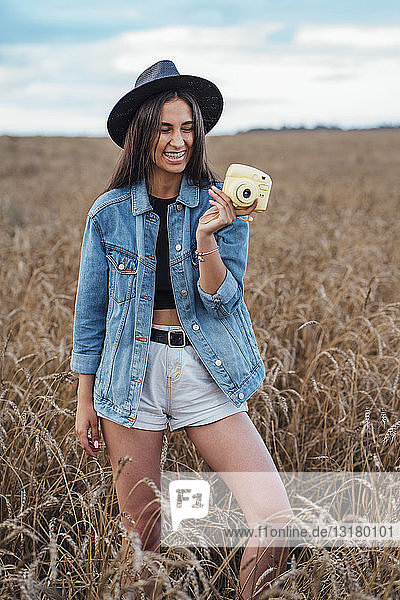 Porträt einer lachenden jungen Frau mit Hut und Jeansjacke  die mit Kamera in einem Maisfeld steht
