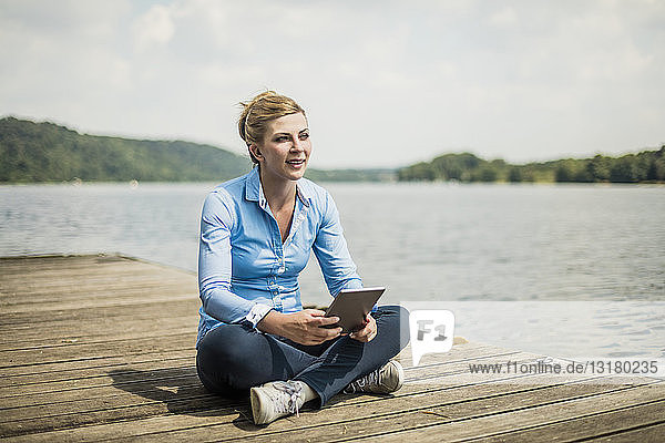 Frau sitzt mit Tablette auf einem Steg an einem See