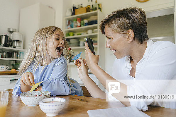 Glückliche Mutter und Tochter amüsieren sich zu Hause am Tisch und fotografieren mit dem Smartphone