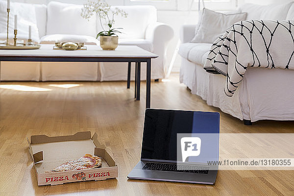 Pizzakarton mit Pizza und Laptop auf dem Boden des Wohnzimmers stehend