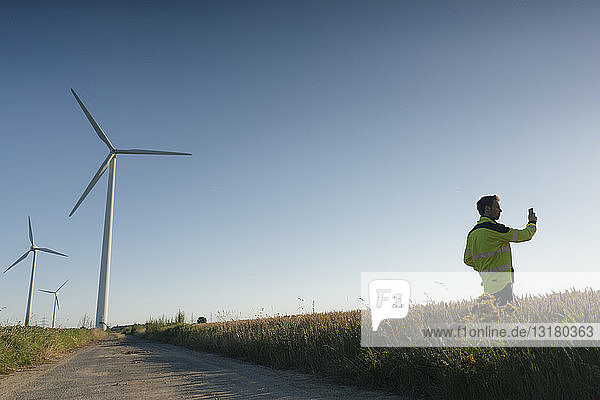 Ingenieur  der in einem Feld bei einem Windpark steht und ein Mobiltelefon benutzt