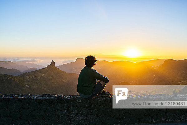 Spanien  Kanarische Inseln  Gran Canaria  Rückenansicht eines Mannes  der auf einer Mauer sitzt und den Sonnenuntergang über einer Berglandschaft betrachtet