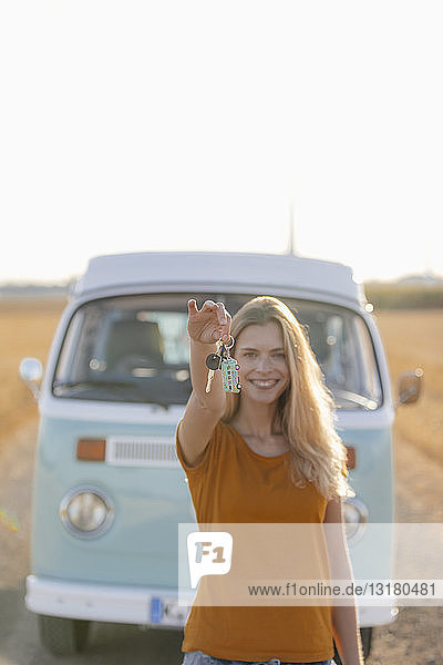 Happy young woman holding car key at camper van in rural landscape
