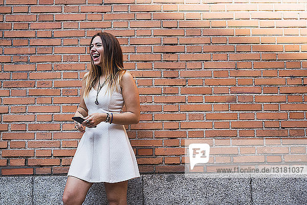 Laughing young woman in front of a brick wall using smartphone