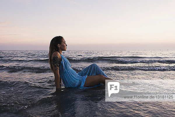 Junge tätowierte Frau in blauem Kleid sitzt am Meeresufer im Wasser und betrachtet Sonnenuntergang