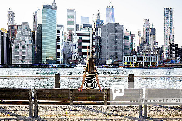 USA  New York  Brooklyn  Rückenansicht einer Frau  die auf einer Bank vor dem East River und der Skyline von Manhattan sitzt