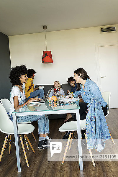 Women using laptop and notebook at dining table with friends in background