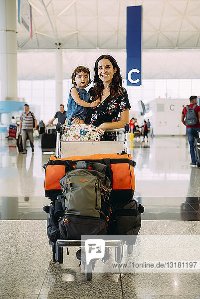 Mutter hält ein kleines Mädchen am Flughafen und schiebt einen Trolley mit dem Gepäck