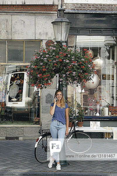 Niederlande  Maastricht  lächelnde junge Frau am Mobiltelefon am Lampenmast in der Stadt