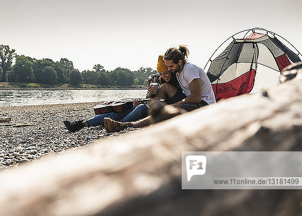 Glückliches junges Paar mit Gitarre sitzt in einem Zelt am Flussufer
