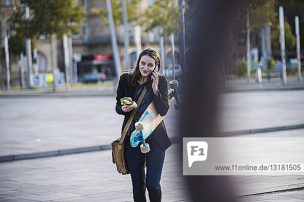 Young woman with longboard and snack in the city on the phone