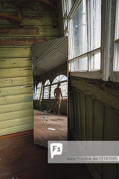 Mirror image of naked man standing at window of abandoned house