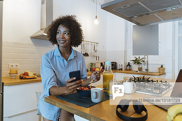 Frau frühstückt in ihrer Küche und benutzt ein Smartphone