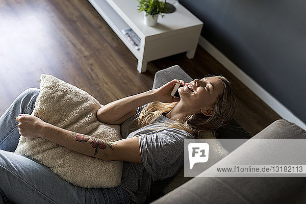 Lächelnde junge Frau mit geschlossenen Augen liegt auf der Couch und telefoniert mit dem Handy
