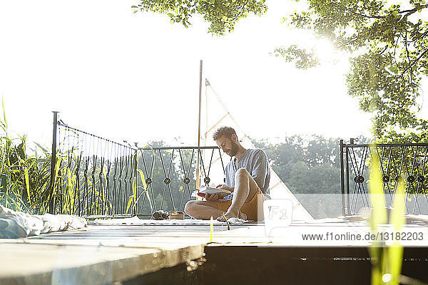 Junger Mann liest Buch auf einem Steg neben einem Segelboot