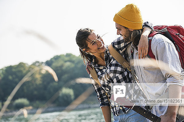 Glückliches junges Paar mit Rucksäcken und Gitarre bei der Umarmung am Flussufer