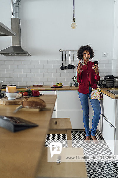 Woman drinking white wine in her kitchen  using smartphone