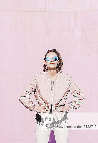Porträt einer jungen Frau mit Sonnenbrille  die vor einer rosa Wand posiert