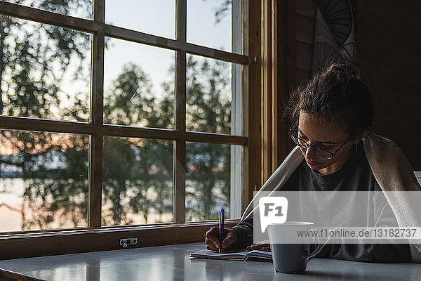 Finnland  Lappland  junge Frau sitzt am Fenster an einem See und schreibt in ihr Tagebuch