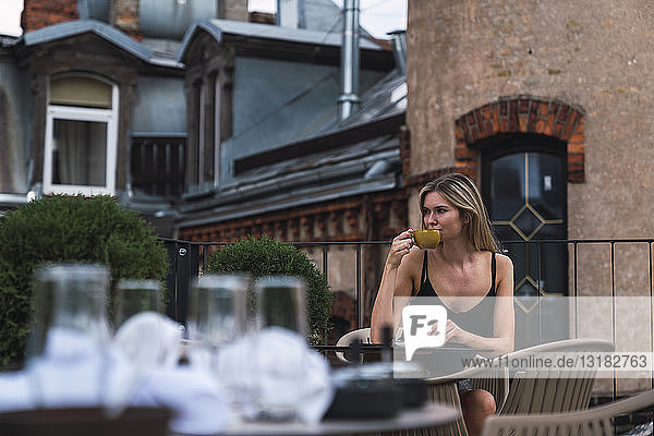 Junge Frau sitzt auf der Dachterrasse und trinkt eine Tasse Kaffee