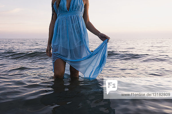 Junge tätowierte Frau in blauem Kleid steht bei Sonnenuntergang im Meer  Teilansicht