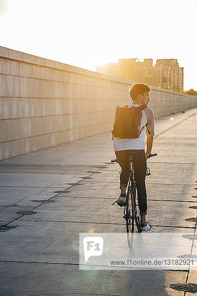 Junger Mann mit Rucksack fährt Fahrrad auf der Promenade bei Sonnenuntergang